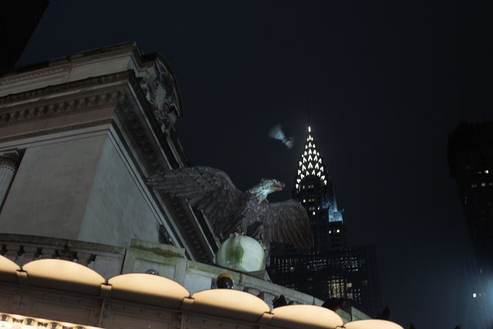 A stone eagle on the Grand Central cornice with the lit Chrysler Building crown behind it.